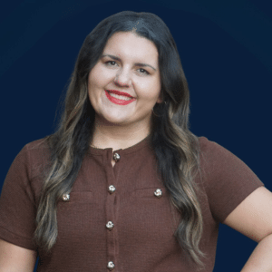 A woman with long, wavy brown hair, wearing a brown short-sleeve top with gold buttons, smiles in front of a dark blue background.