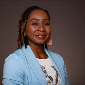 A woman with braided hair, wearing a light blue blazer, patterned white top, and colorful earrings, smiles in front of a plain brown background.