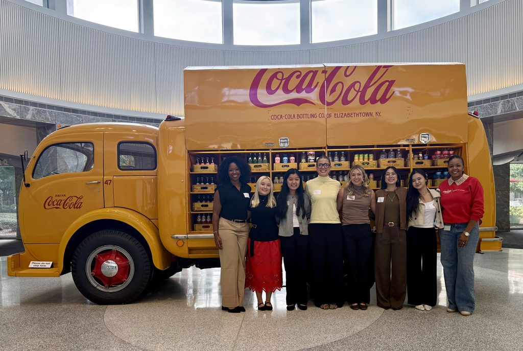 Eight women stand in front of a vintage yellow Coca-Cola delivery truck inside a building.
