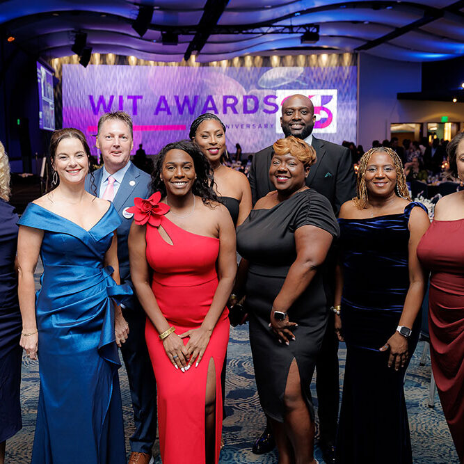 Nine people dressed formally pose and smile at the WIT Awards event. The background shows event signage, stage lighting, and seated guests in a large, elegant venue.