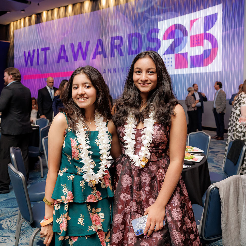 Two young women wearing floral dresses and leis smile at a formal event. Behind them is a large lit screen that reads WIT AWARDS 25. Other attendees and tables are visible in the background.
